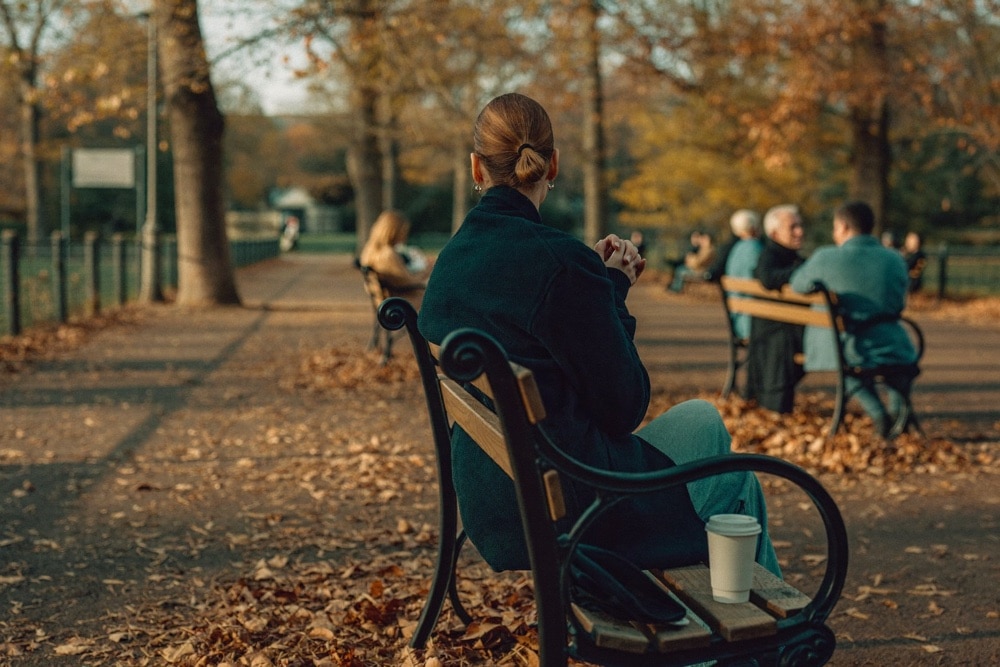 Person sitting alone on a park bench in autumn light, groups talking on benches nearby